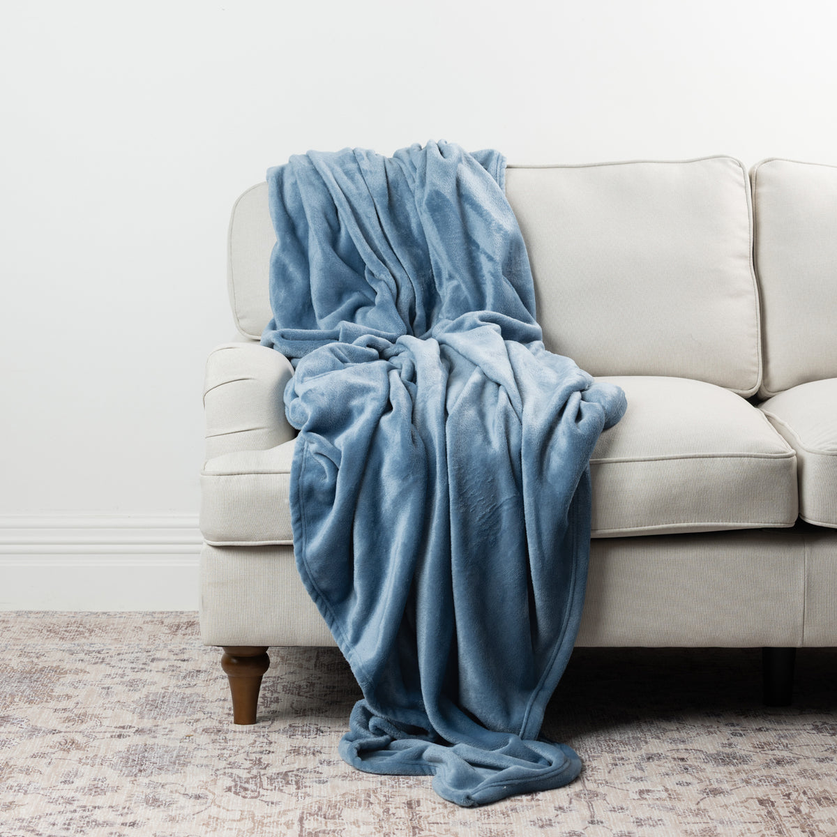 Faded blue Home Co. Super Plush Blanket draped over a neutral sofa, highlighting its smooth plush texture and relaxed drape.