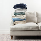 Stack of coordinating Home Co. Super Plush Blankets in navy, silver, pine green, natural, faded blue and champagne displayed on a neutral sofa.