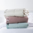 Fieldcrest Duck Down Blankets in Natural, Blush and Duckegg, shown rolled and stacked together on a white bed.