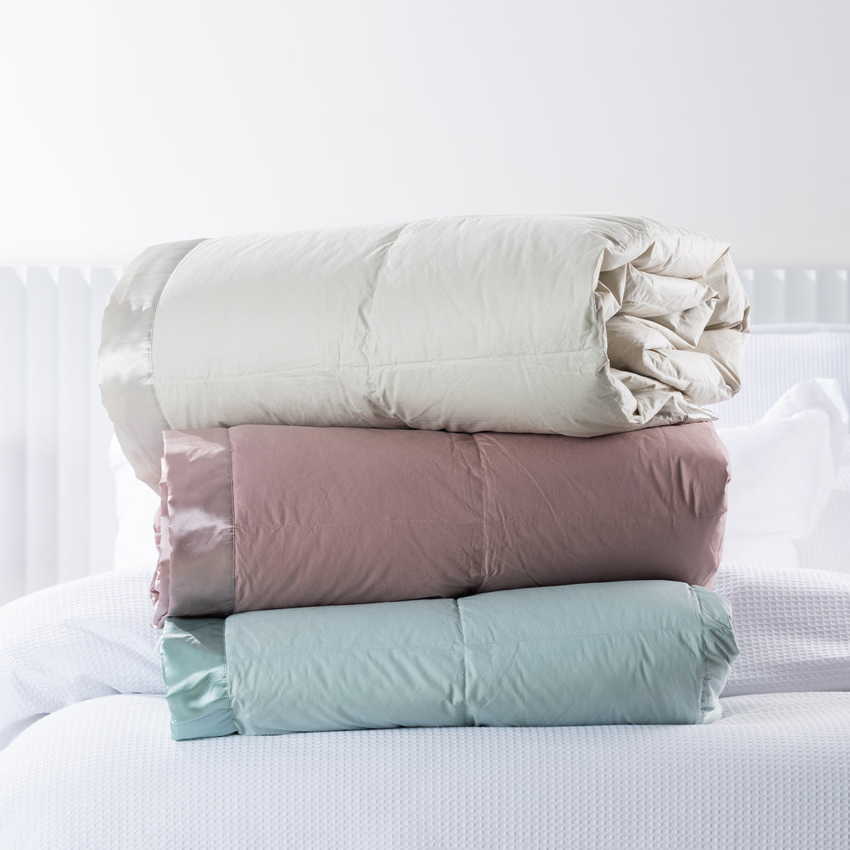 Fieldcrest Duck Down Blankets in Natural, Blush and Duckegg, shown rolled and stacked together on a white bed.