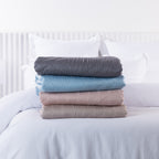 Bambou Summer Blanket range folded and stacked on a white bed, showing Charcoal, Blue, Blush and Mushroom colour options.