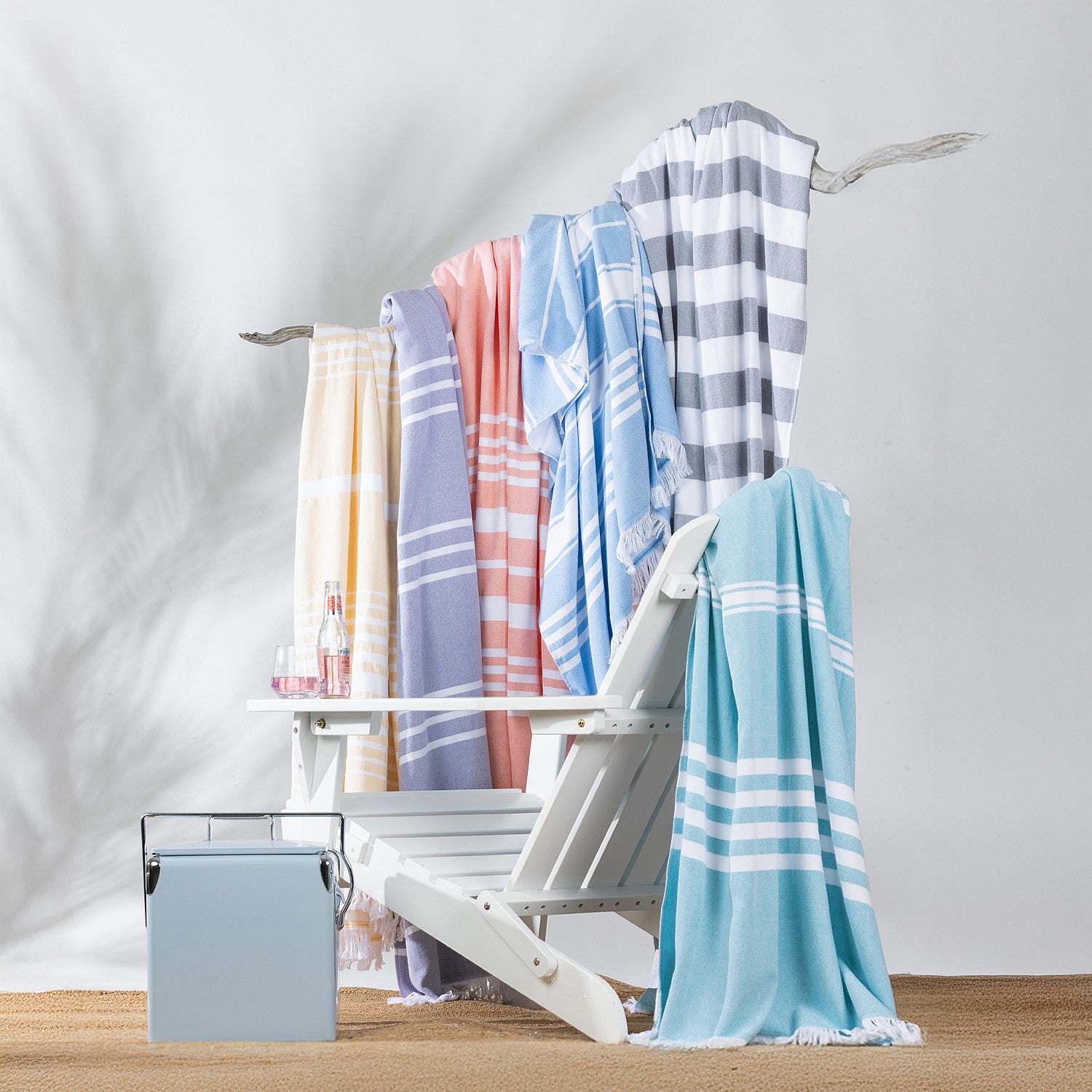 Collection of striped beach towels in assorted pastel and neutral colours draped over a white outdoor lounge chair in a relaxed summer setting.