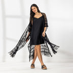 A woman modeling a black slip dress with a sheer black lace kaftan cover-up draped over her, set against a bright white studio background.