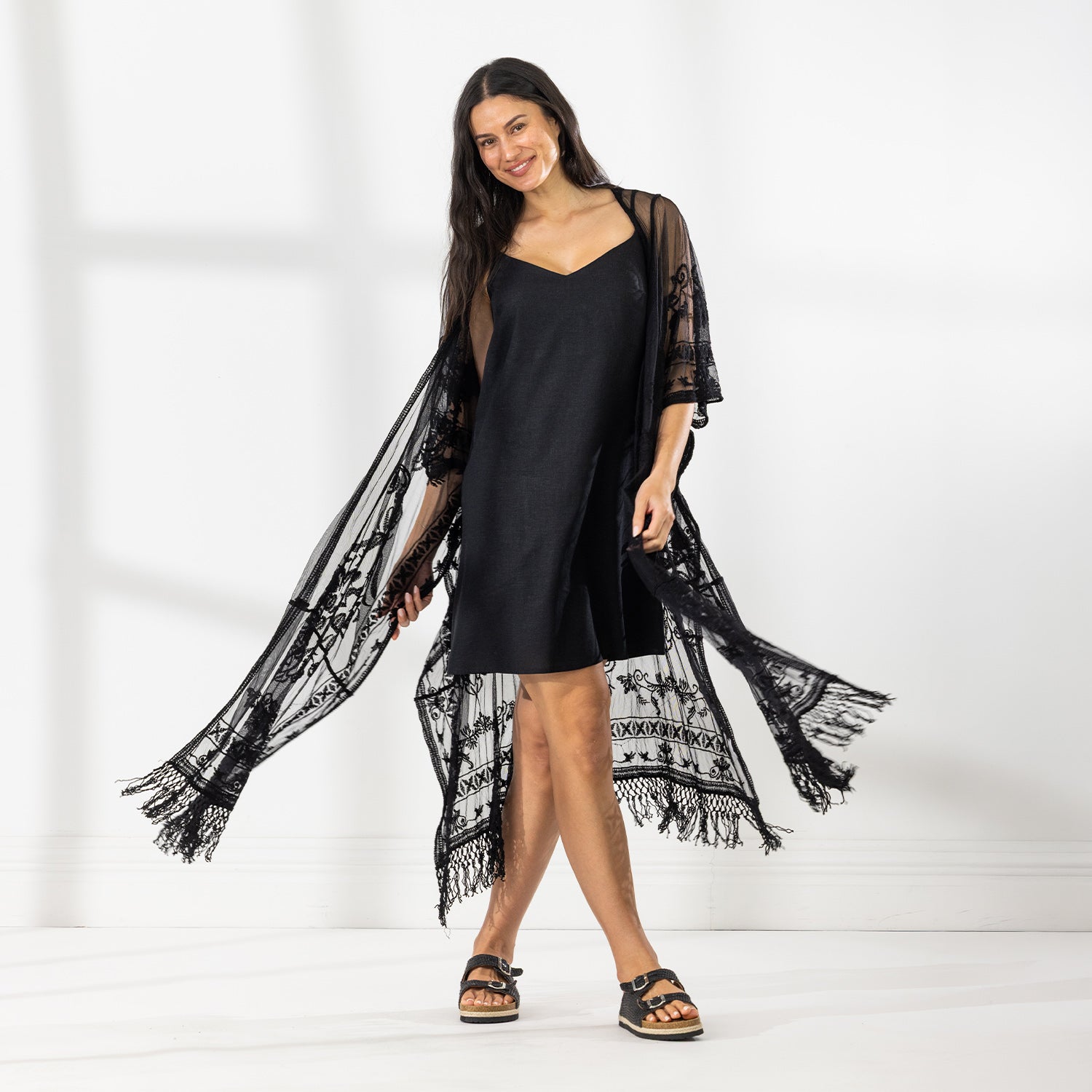 A woman modeling a black slip dress with a sheer black lace kaftan cover-up draped over her, set against a bright white studio background.