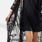 A woman modeling a black slip dress with a sheer black lace kaftan cover-up draped over her, set against a bright white studio background.