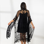 A woman modeling a black slip dress with a sheer black lace kaftan cover-up draped over her, set against a bright white studio background.