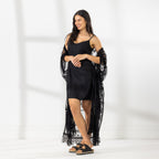 A woman modeling a black slip dress with a sheer black lace kaftan cover-up draped over her, set against a bright white studio background.