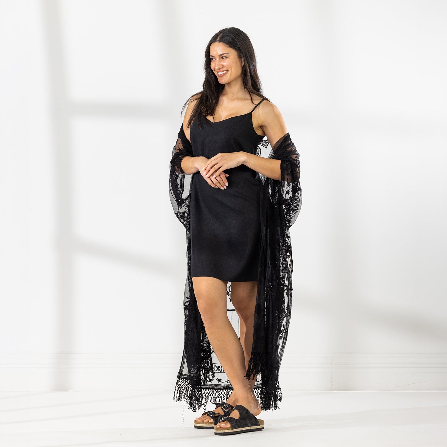 A woman modeling a black slip dress with a sheer black lace kaftan cover-up draped over her, set against a bright white studio background.
