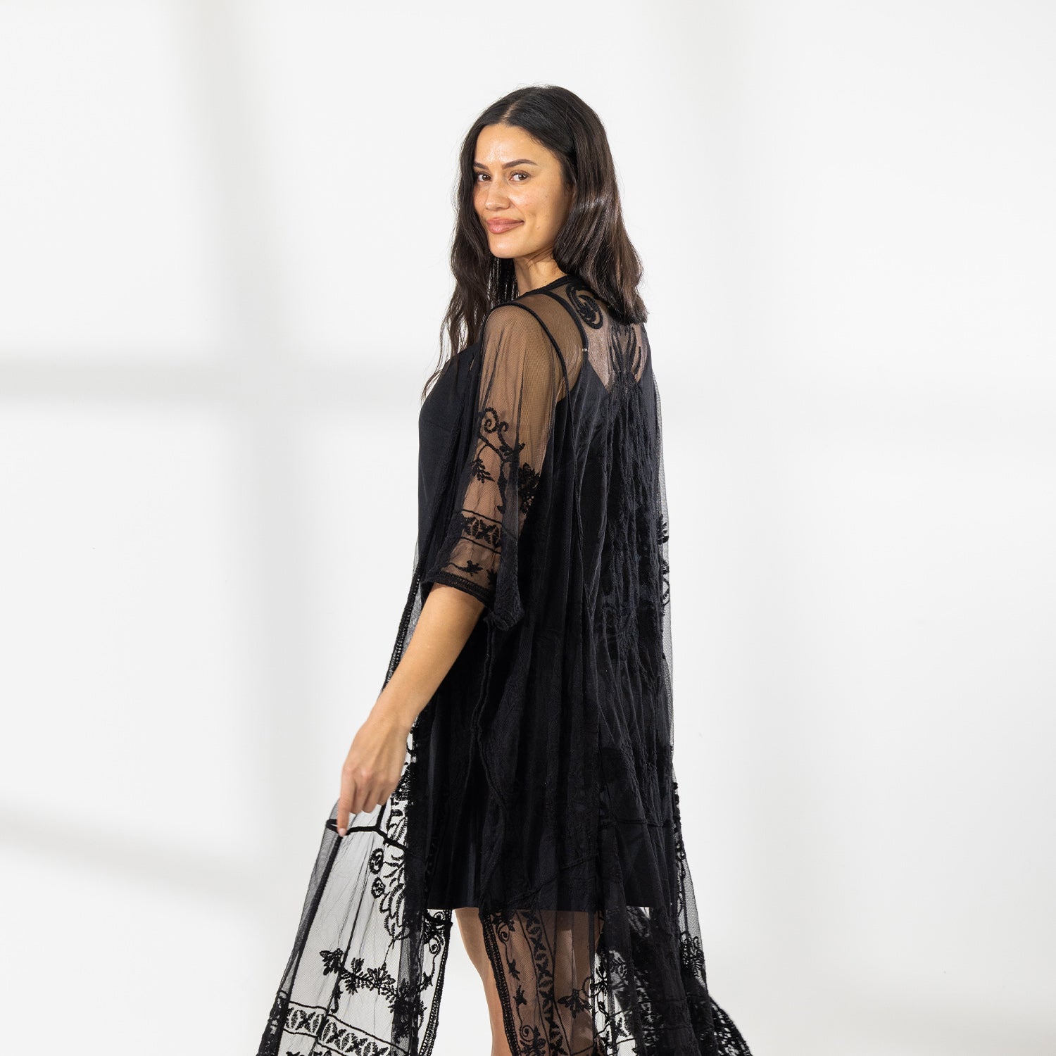 A woman modeling a black slip dress with a sheer black lace kaftan cover-up draped over her, set against a bright white studio background.