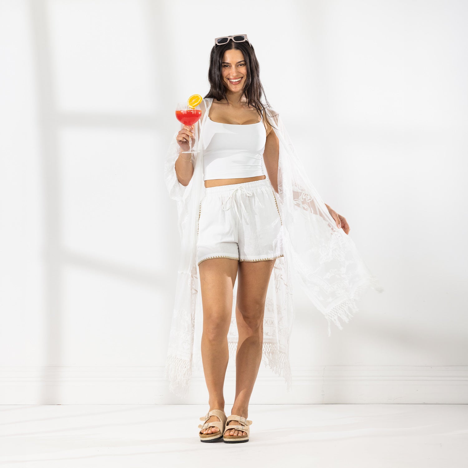 A woman stands in a bright white studio wearing a white two-piece crop top and shorts set with a sheer white fringed cover-up draped over her arms. She is holding a pink cocktail with an orange slice garnish and wearing beige sandals.