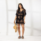 A woman modeling a black sheer lace cover-up dress with fringe, holding a straw hat against a bright white studio background.