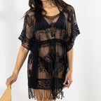 A woman modeling a black sheer lace cover-up dress with fringe, holding a straw hat against a bright white studio background.