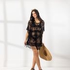 A woman modeling a black sheer lace cover-up dress with fringe, holding a straw hat against a bright white studio background.