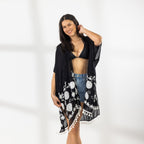 A woman modeling a black open-front kaftan cover-up with white Floral patterns and fringe trim, worn over a black bikini top and blue denim shorts in a bright studio setting.