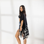 A woman modeling a black open-front kaftan cover-up with white Floral patterns and fringe trim, worn over a black bikini top and blue denim shorts in a bright studio setting.