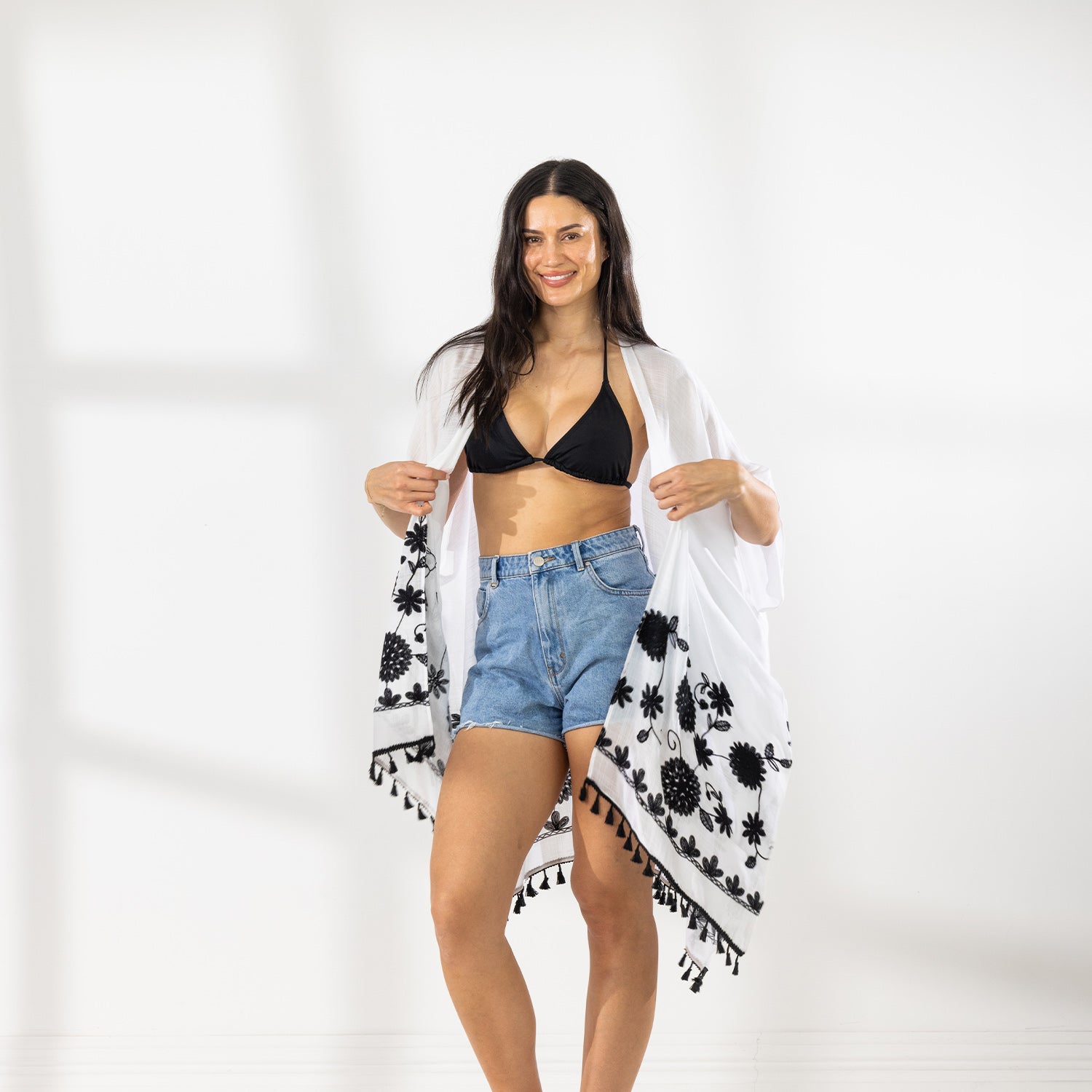 A woman modeling a White open-front kaftan cover-up with white Floral patterns and fringe trim, worn over a black bikini top and blue denim shorts in a bright studio setting.
