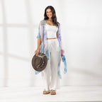 A woman stands in a bright white studio wearing a sheer pastel tie-dye kimono draped over a white crop top and white pants, while holding a brown woven hat.