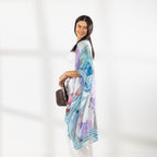 A woman modeling a lightweight open-front kaftan with a blue, purple, and pink watercolor-style print, worn over a white crop top and white drawstring pants while holding a brown hat against a bright white background.