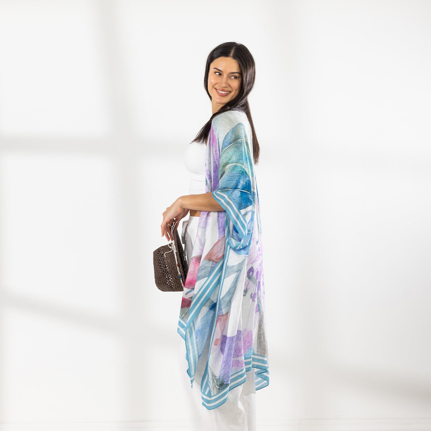 A woman modeling a lightweight open-front kaftan with a blue, purple, and pink watercolor-style print, worn over a white crop top and white drawstring pants while holding a brown hat against a bright white background.