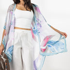 A woman modeling a lightweight open-front kaftan with a blue, purple, and pink watercolor-style print, worn over a white crop top and white drawstring pants while holding a brown hat against a bright white background.