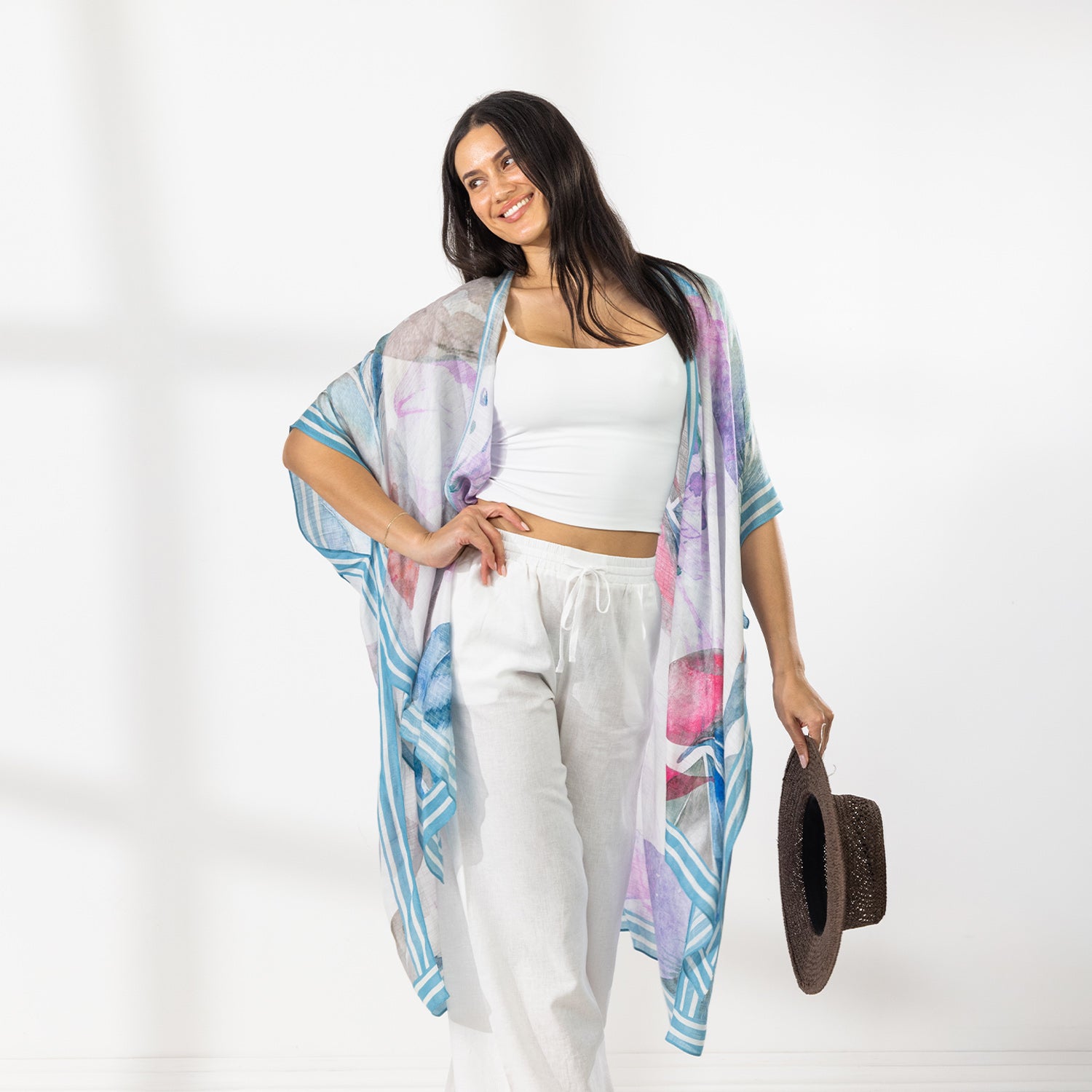 A woman modeling a lightweight open-front kaftan with a blue, purple, and pink watercolor-style print, worn over a white crop top and white drawstring pants while holding a brown hat against a bright white background.