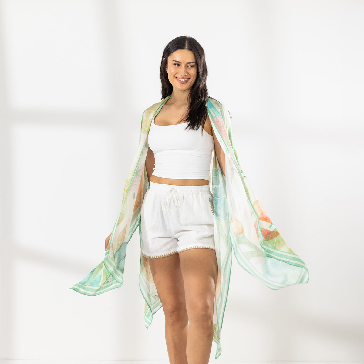A woman stands in a bright white studio wearing a white two-piece outfit with white shorts, tan sandals, and a sheer green and white patterned Kaftan draped over her shoulders.