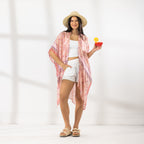 A woman stands in a bright white studio wearing a light pink patterned open-front kaftan cover-up over a white crop top and white shorts, with a straw hat and sandals. She holds a red cocktail in one hand.