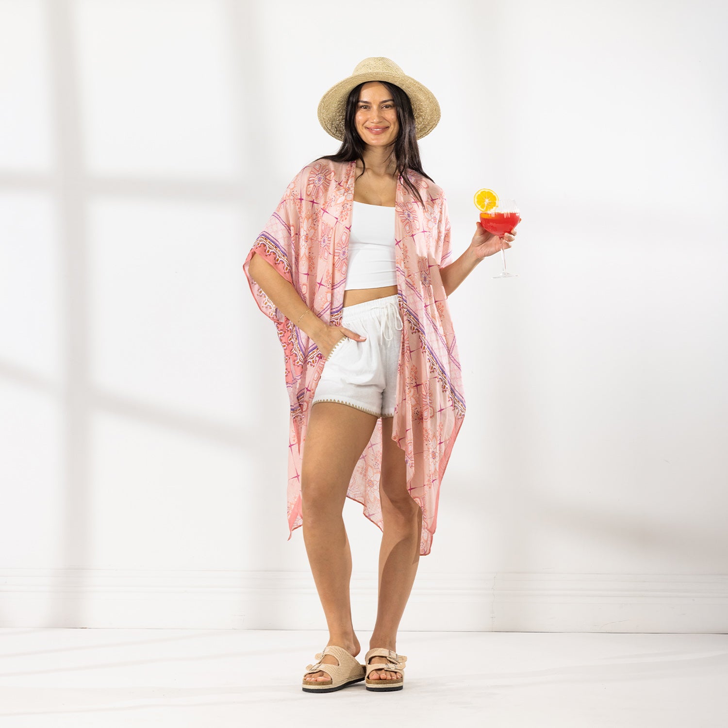 A woman stands in a bright white studio wearing a light pink patterned open-front kaftan cover-up over a white crop top and white shorts, with a straw hat and sandals. She holds a red cocktail in one hand.