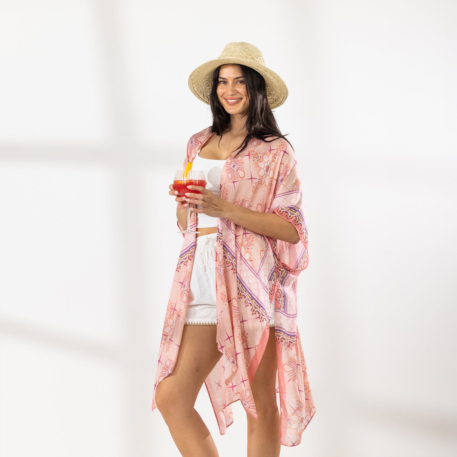 A woman stands in a bright white studio wearing a light pink patterned open-front kaftan cover-up over a white crop top and white shorts, with a straw hat and sandals. She holds a red cocktail in one hand.