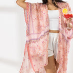 A woman stands in a bright white studio wearing a light pink patterned open-front kaftan cover-up over a white crop top and white shorts, with a straw hat and sandals. She holds a red cocktail in one hand.