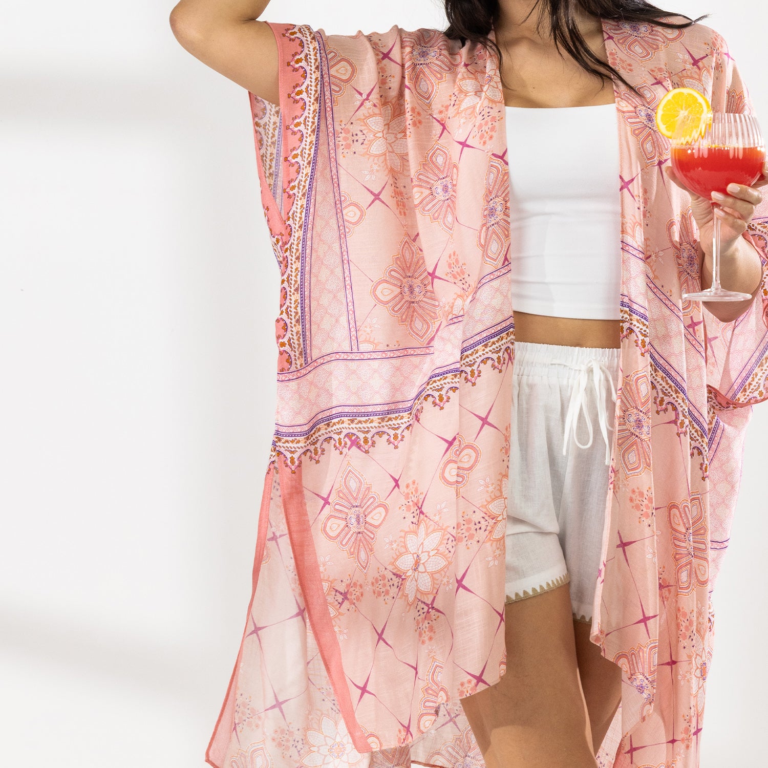 A woman stands in a bright white studio wearing a light pink patterned open-front kaftan cover-up over a white crop top and white shorts, with a straw hat and sandals. She holds a red cocktail in one hand.