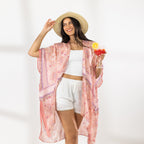 A woman stands in a bright white studio wearing a light pink patterned open-front kaftan cover-up over a white crop top and white shorts, with a straw hat and sandals. She holds a red cocktail in one hand.