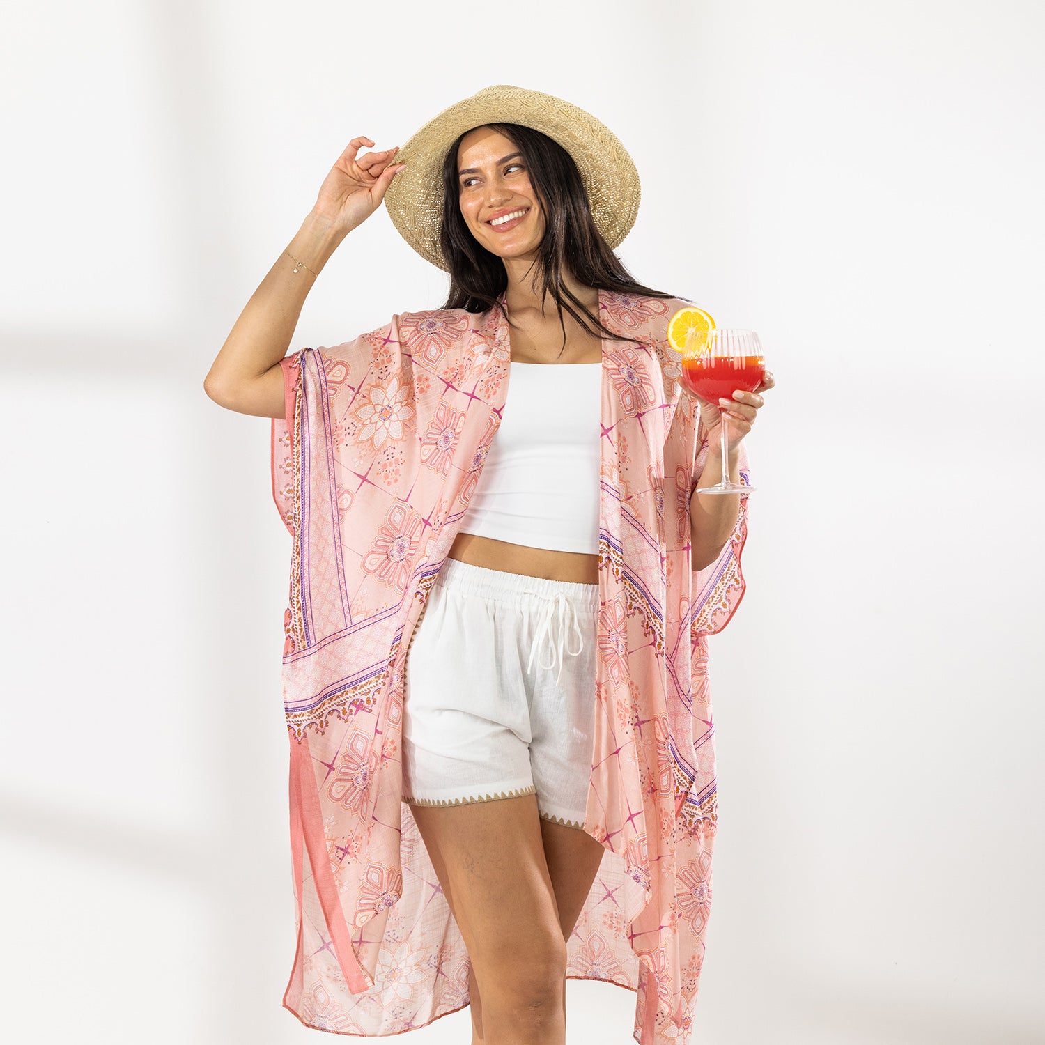 A woman stands in a bright white studio wearing a light pink patterned open-front kaftan cover-up over a white crop top and white shorts, with a straw hat and sandals. She holds a red cocktail in one hand.