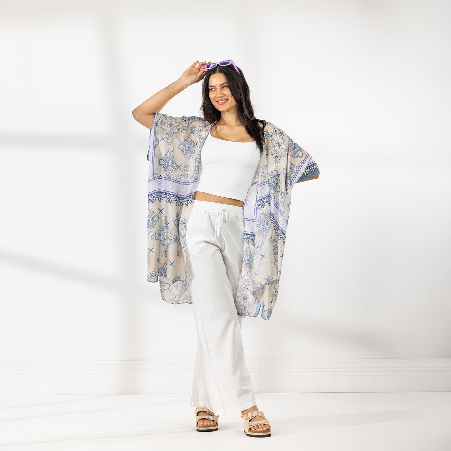 A woman modeling a lightweight open-front kaftan with a beige base and blue floral bohemian pattern, styled over a white crop top and white wide-leg pants.