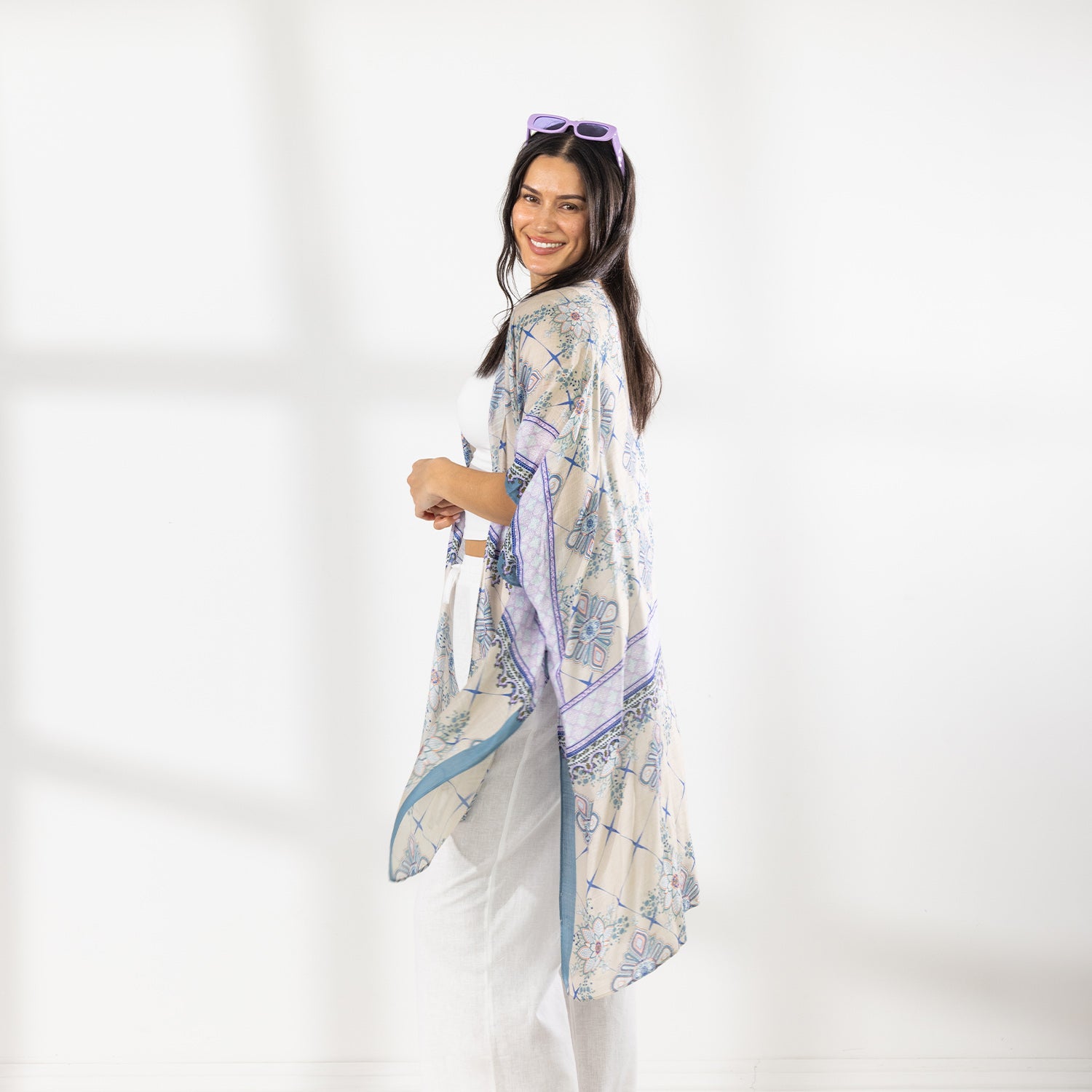 A woman modeling a lightweight open-front kaftan with a beige base and blue floral bohemian pattern, styled over a white crop top and white wide-leg pants.