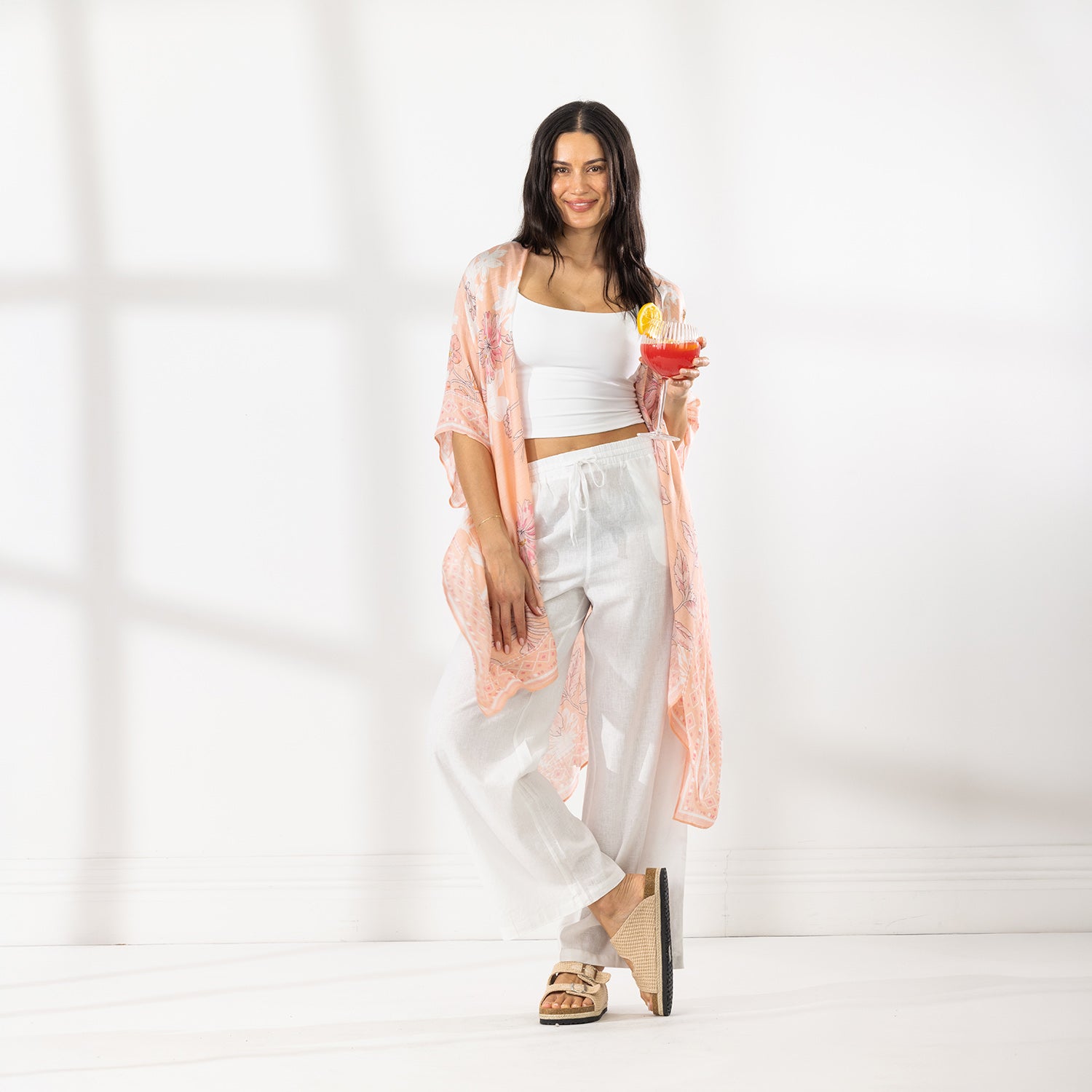 A woman stands in a bright white room wearing a white crop top, white wide-leg pants, and a sheer peach floral kaftan, holding a colorful cocktail.