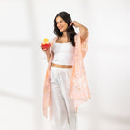 A woman stands in a bright white room wearing a white crop top, white wide-leg pants, and a sheer peach floral kaftan, holding a colorful cocktail.
