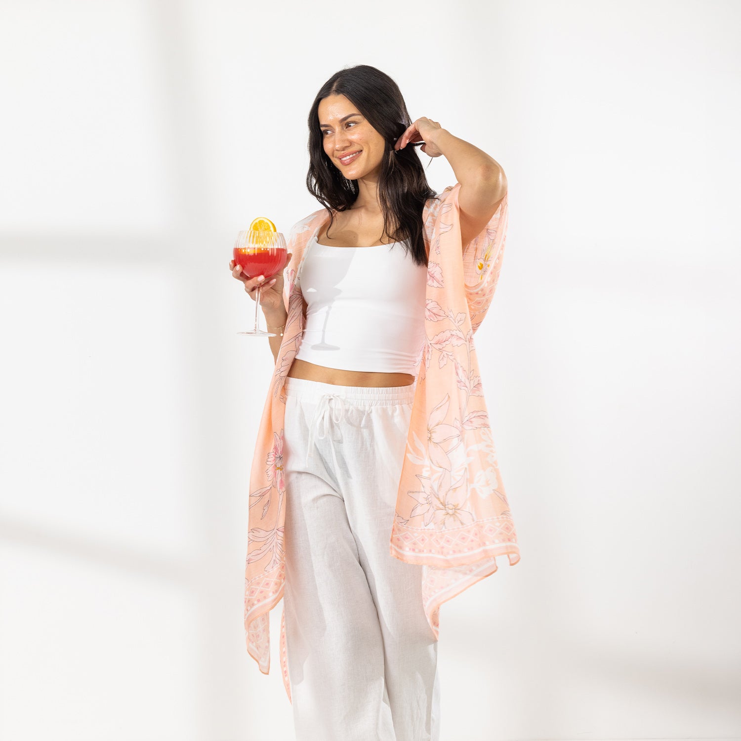 A woman stands in a bright white room wearing a white crop top, white wide-leg pants, and a sheer peach floral kaftan, holding a colorful cocktail.
