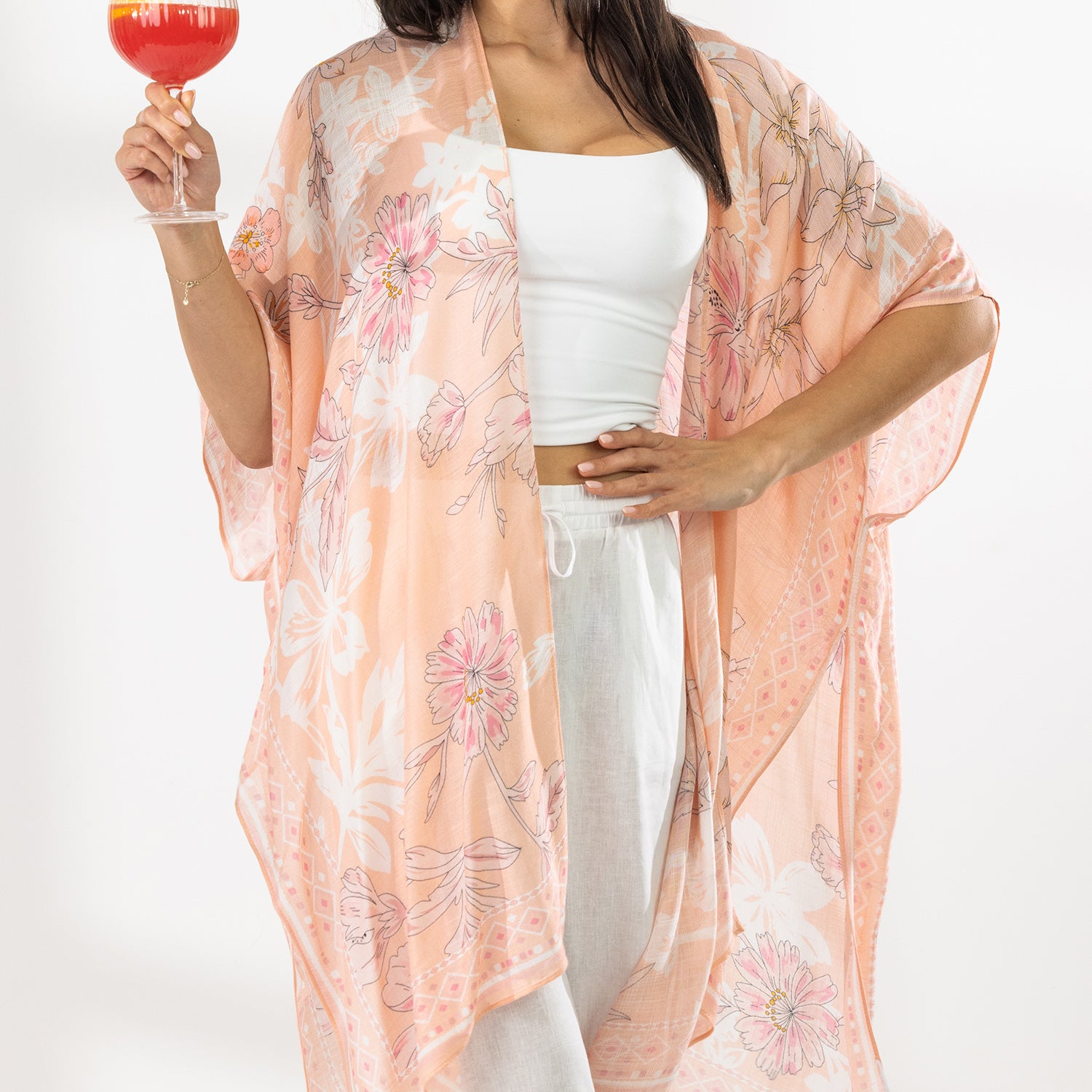 A woman stands in a bright white room wearing a white crop top, white wide-leg pants, and a sheer peach floral kaftan, holding a colorful cocktail.