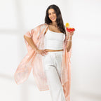 A woman stands in a bright white room wearing a white crop top, white wide-leg pants, and a sheer peach floral kaftan, holding a colorful cocktail.
