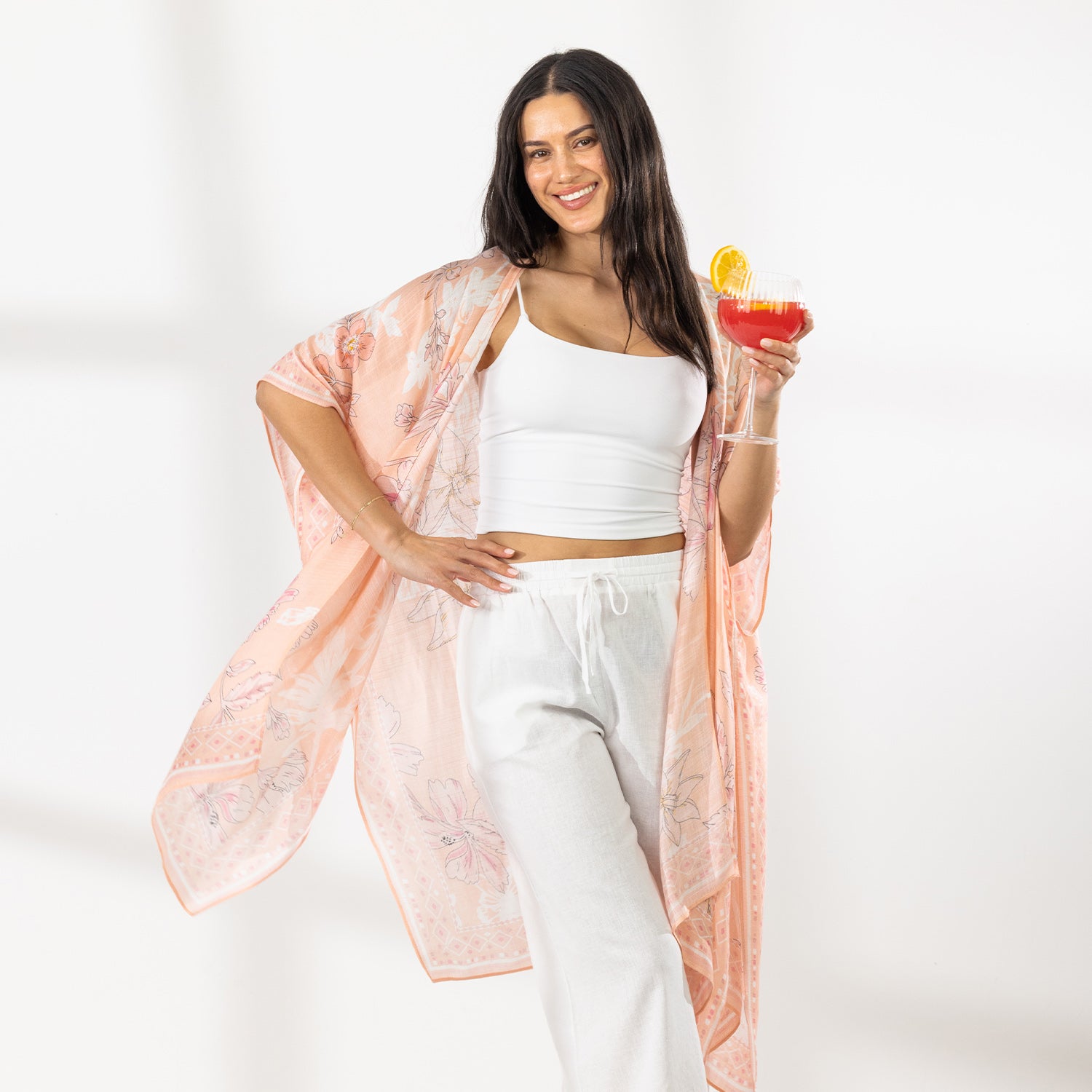 A woman stands in a bright white room wearing a white crop top, white wide-leg pants, and a sheer peach floral kaftan, holding a colorful cocktail.