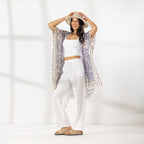 A woman stands in a bright white studio wearing a white cropped tank top, white wide-leg pants, beige sandals, and a beige wide-brim hat. She has a light purple and cream patterned open-front kaftan draped over her shoulders.