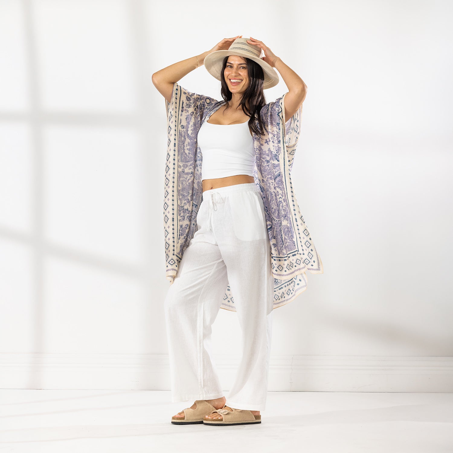 A woman stands in a bright white studio wearing a white cropped tank top, white wide-leg pants, beige sandals, and a beige wide-brim hat. She has a light purple and cream patterned open-front kaftan draped over her shoulders.