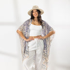 A woman stands in a bright white studio wearing a white cropped tank top, white wide-leg pants, beige sandals, and a beige wide-brim hat. She has a light purple and cream patterned open-front kaftan draped over her shoulders.