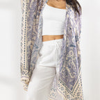 A woman stands in a bright white studio wearing a white cropped tank top, white wide-leg pants, beige sandals, and a beige wide-brim hat. She has a light purple and cream patterned open-front kaftan draped over her shoulders.