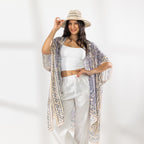 A woman stands in a bright white studio wearing a white cropped tank top, white wide-leg pants, beige sandals, and a beige wide-brim hat. She has a light purple and cream patterned open-front kaftan draped over her shoulders.
