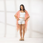 A woman modeling a white crop top and white shorts with a peach and orange patterned open-front kaftan draped over her shoulders, standing against a white wall with soft window shadows.