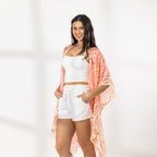 A woman modeling a white crop top and white shorts with a peach and orange patterned open-front kaftan draped over her shoulders, standing against a white wall with soft window shadows.