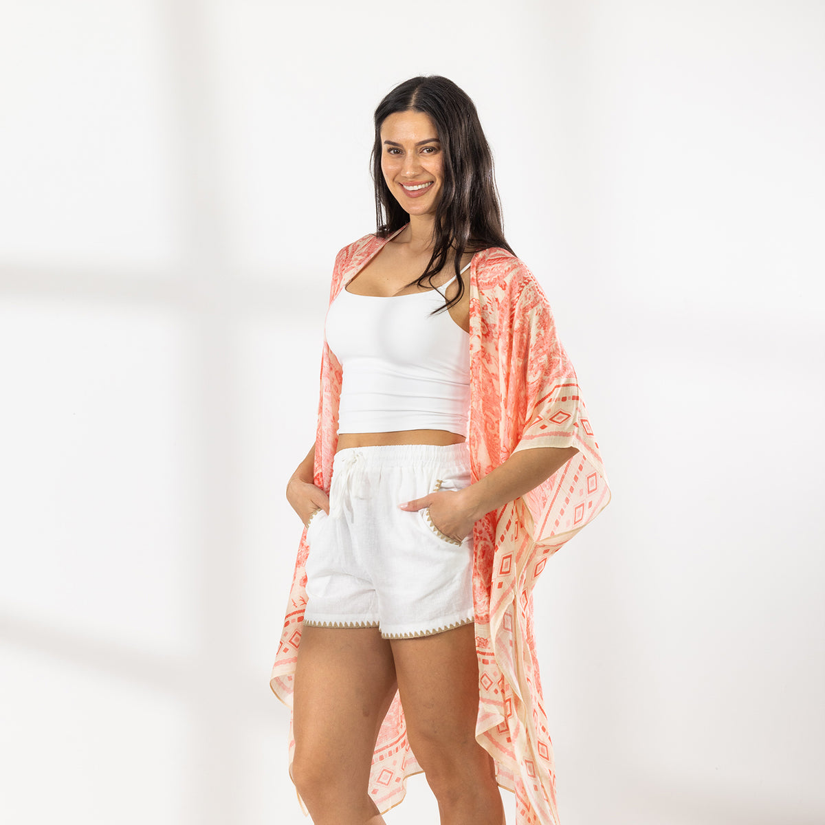 A woman modeling a white crop top and white shorts with a peach and orange patterned open-front kaftan draped over her shoulders, standing against a white wall with soft window shadows.