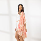 A woman modeling a white crop top and white shorts with a peach and orange patterned open-front kaftan draped over her shoulders, standing against a white wall with soft window shadows.